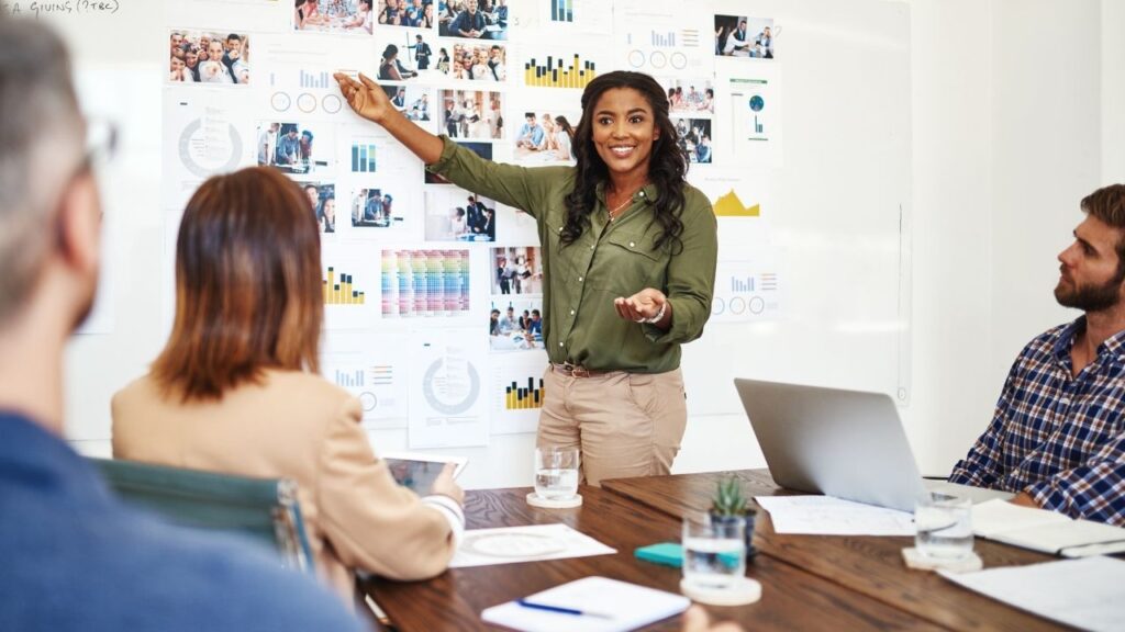 Mulher de bluda verde realizando uma apresentação em uma sala com algumas pessoas utilizando diversos gráficos grudados em um quadra branco na parede.