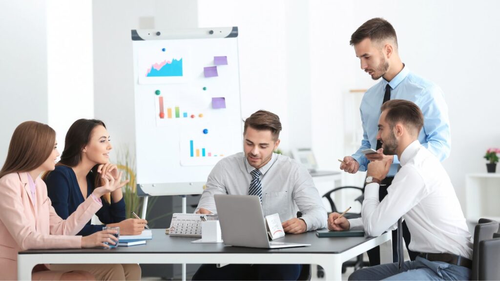 Grupo de 2 mulheres e 3 homens sentados em volta de uma mesa conversando com um quadro com gráficos ao fundo.